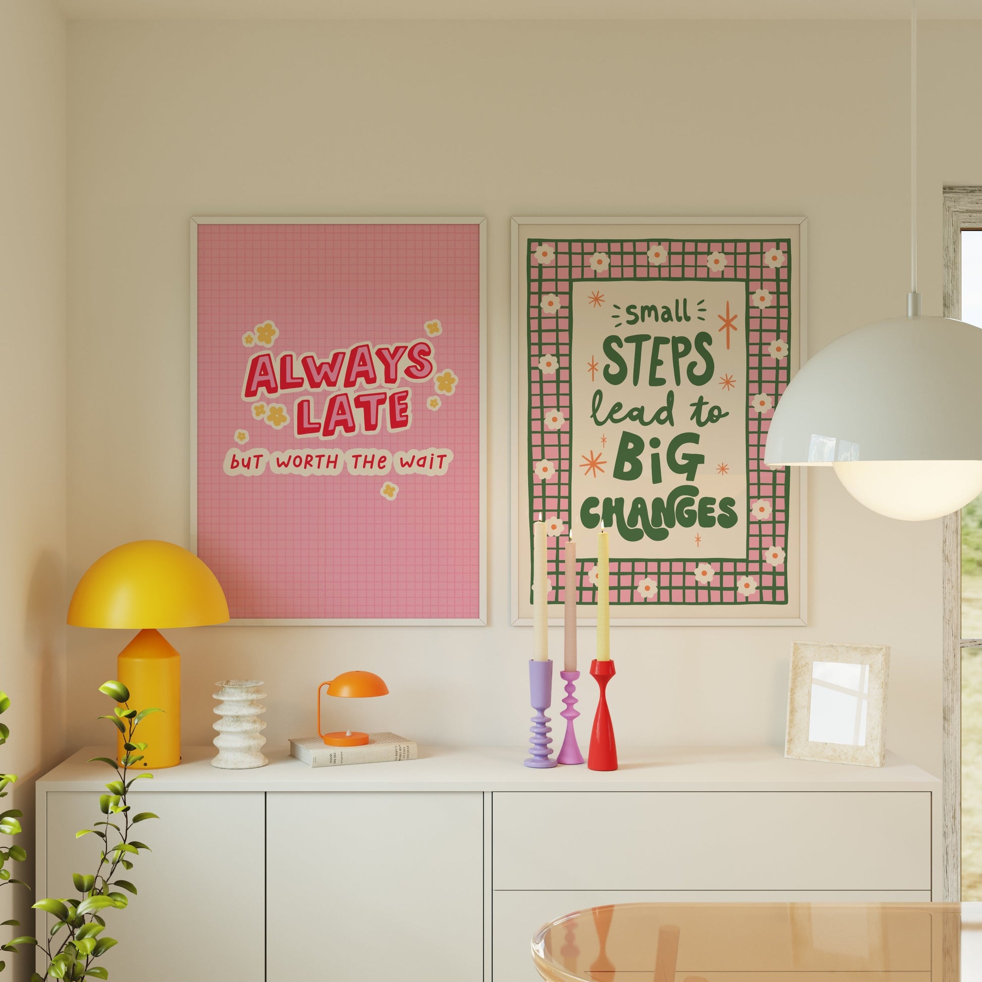 Two framed motivational art prints hanging on a beige wall above a white sideboard in a bright, modern room. The left print is pink with the text 'ALWAYS LATE BUT WORTH THE WAIT' in red and yellow.