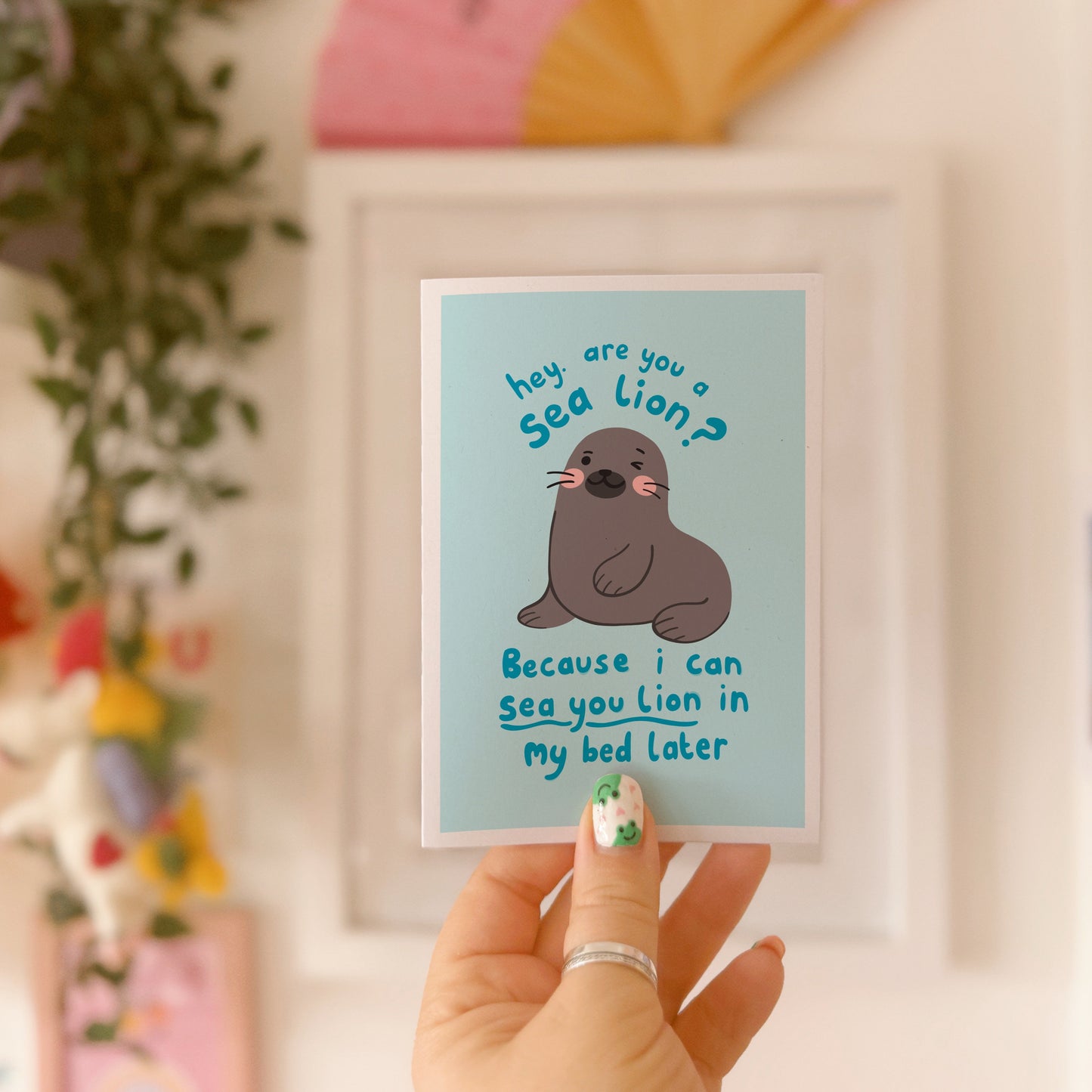 A blue greeting card with an illustration of a sea lion on the front. There is a cheeky quote on the front, using a pun, saying 'are you a sea lion? because I can sea you lion in my bed later'.