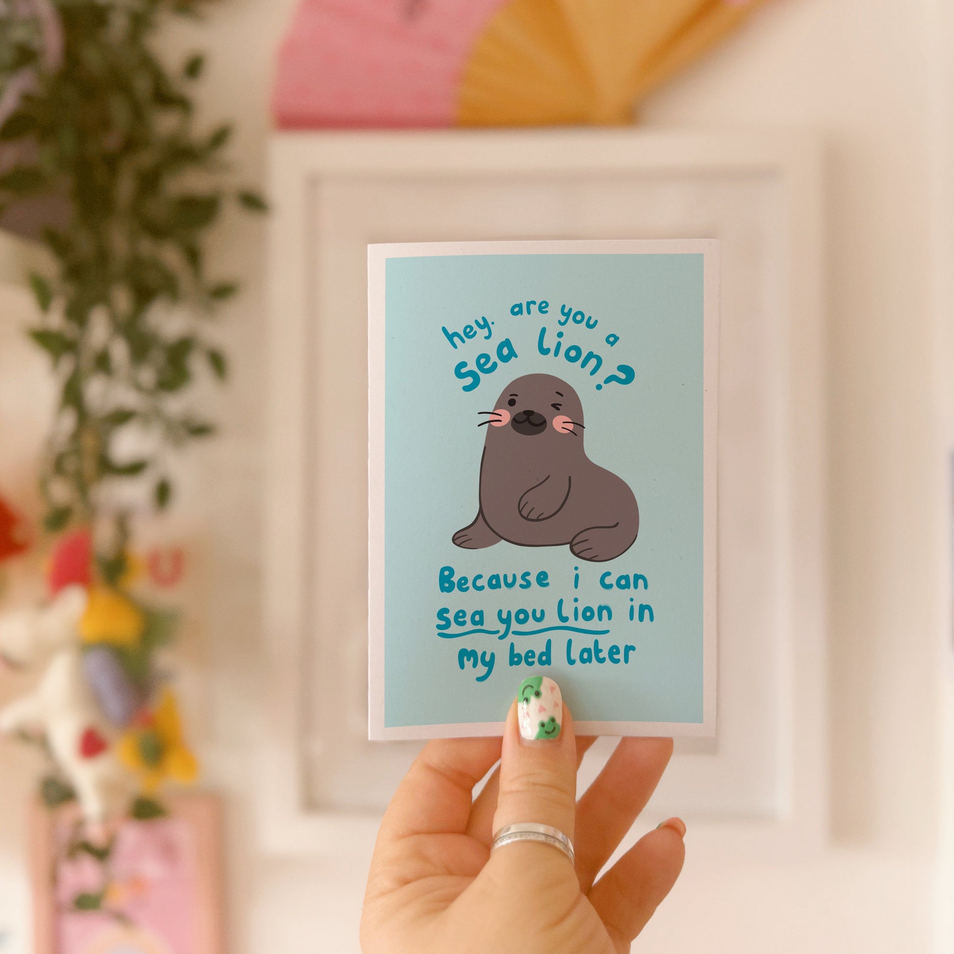 A blue greeting card with an illustration of a sea lion on the front. There is a cheeky quote on the front, using a pun, saying 'are you a sea lion? because I can sea you lion in my bed later'.