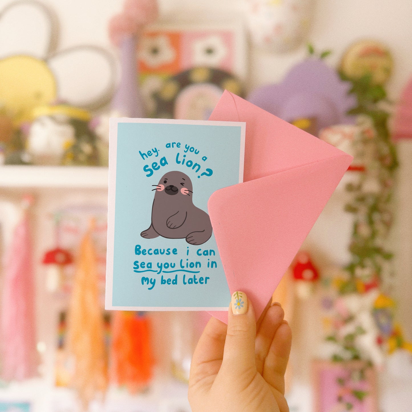 A blue greeting card with an illustration of a sea lion on the front. There is a cheeky quote on the front, using a pun, saying 'are you a sea lion? because I can sea you lion in my bed later'.