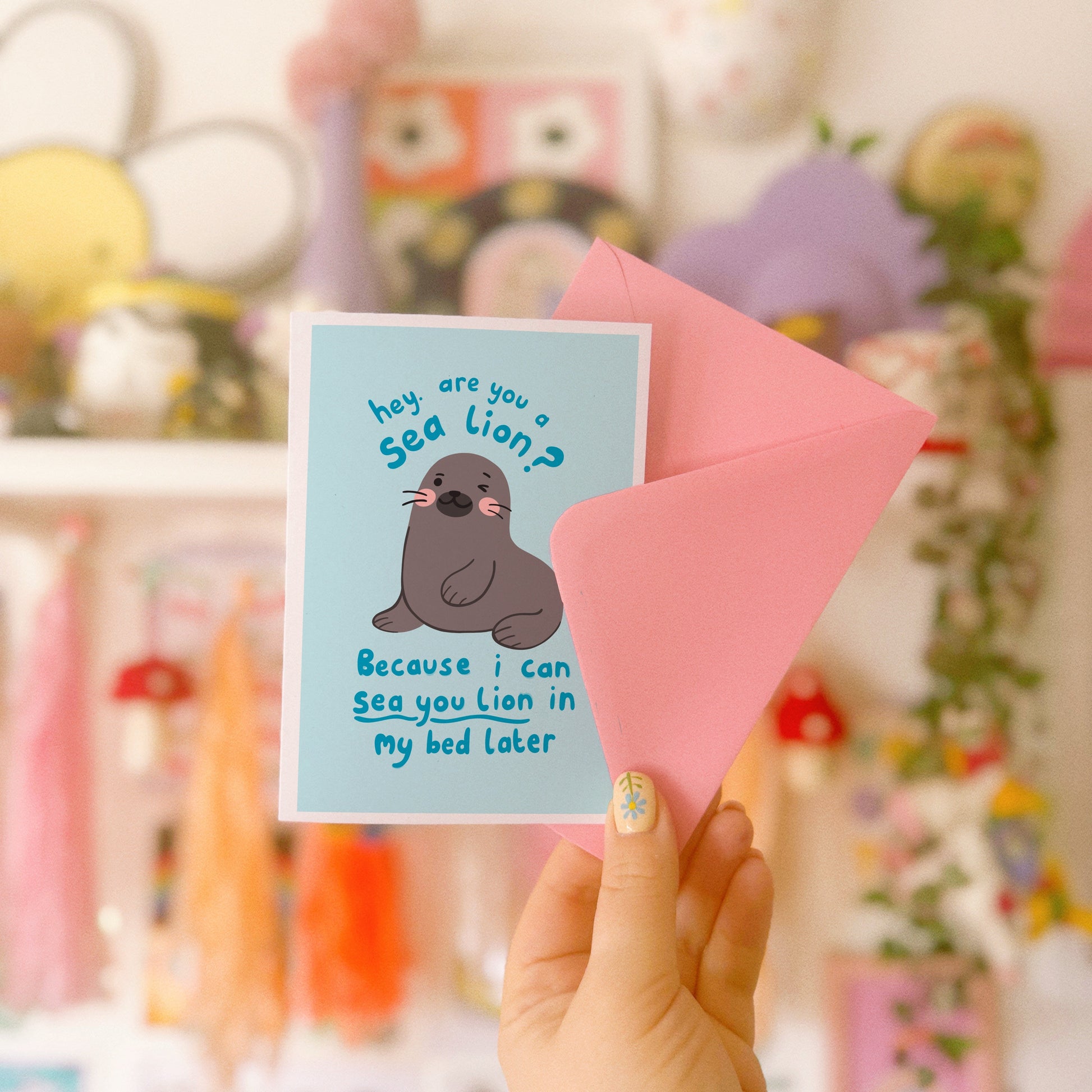 A blue greeting card with an illustration of a sea lion on the front. There is a cheeky quote on the front, using a pun, saying 'are you a sea lion? because I can sea you lion in my bed later'.