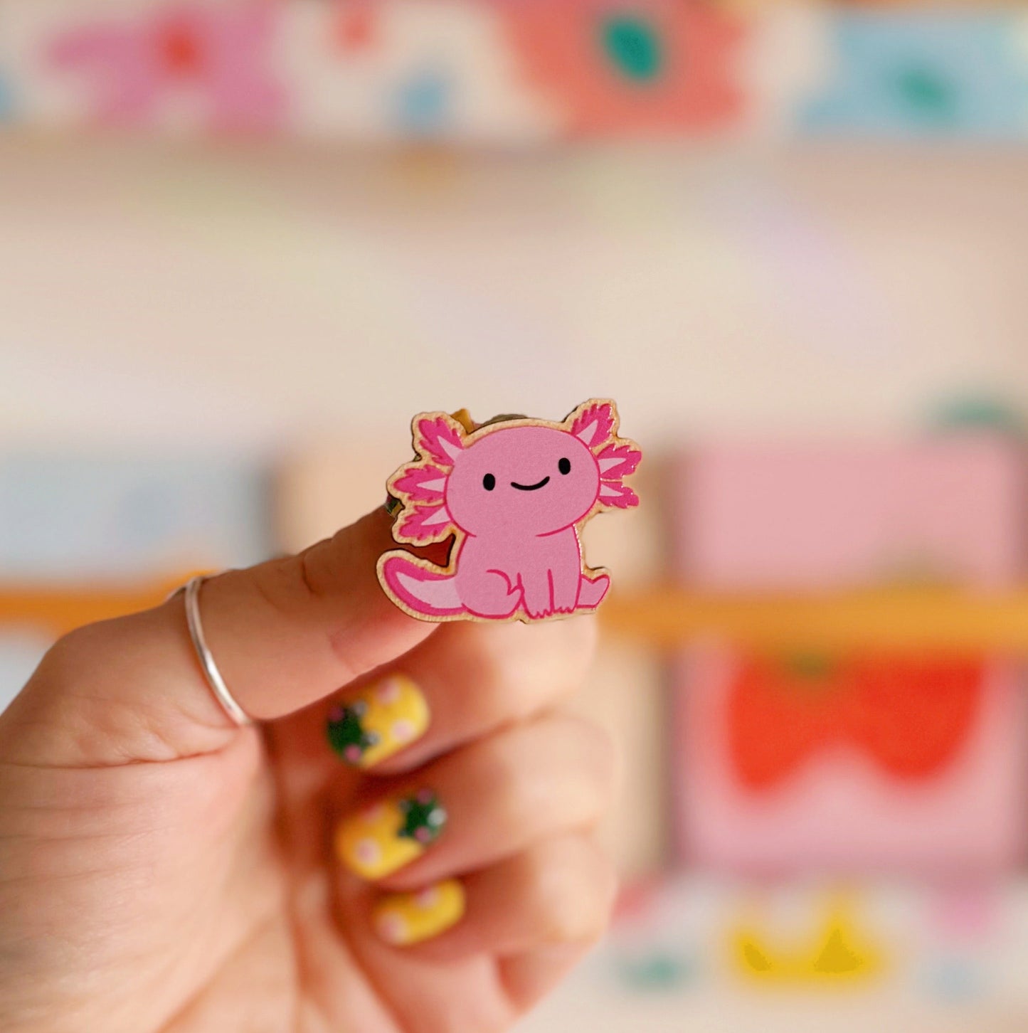 A wooden pin badge with a glossy finish, featuring an illustration of a pink axolotl.