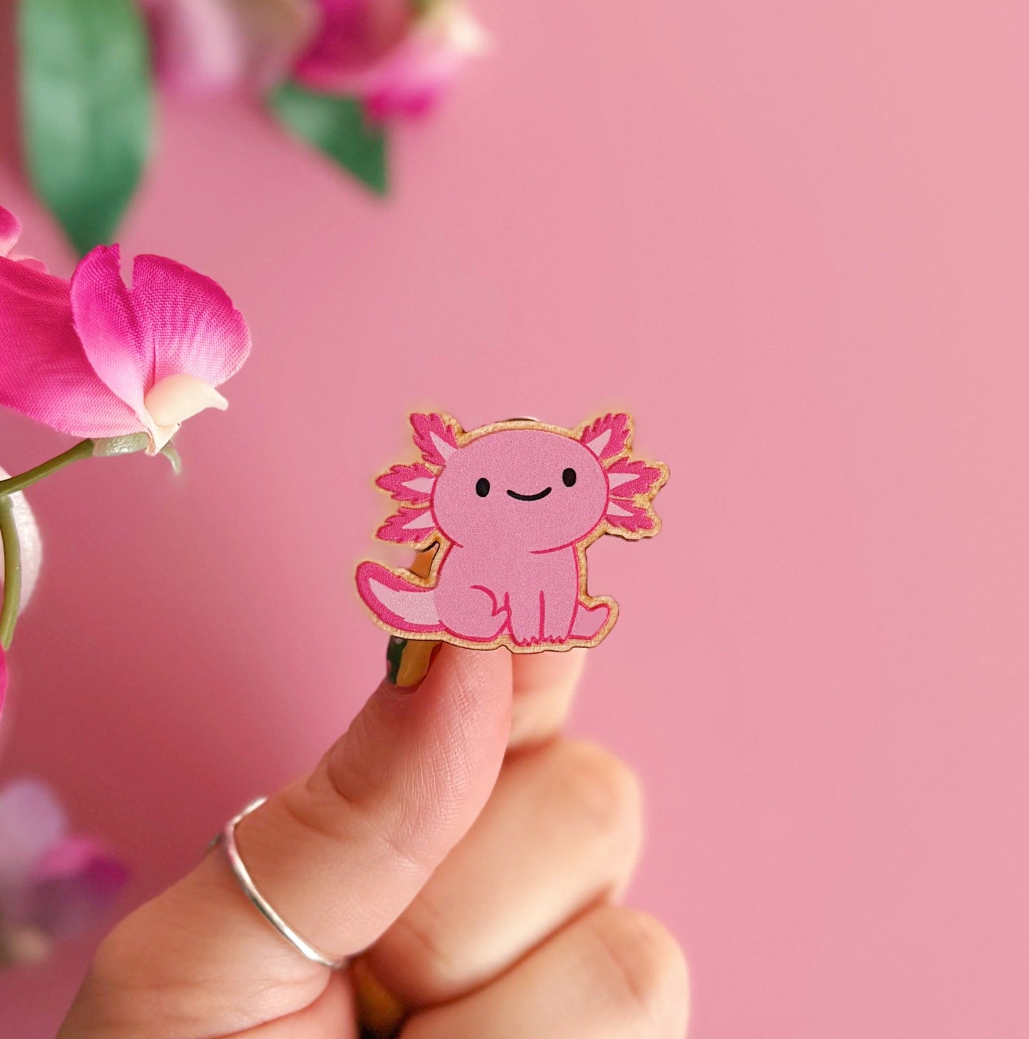 A wooden pin badge with a glossy finish, featuring an illustration of a pink axolotl.