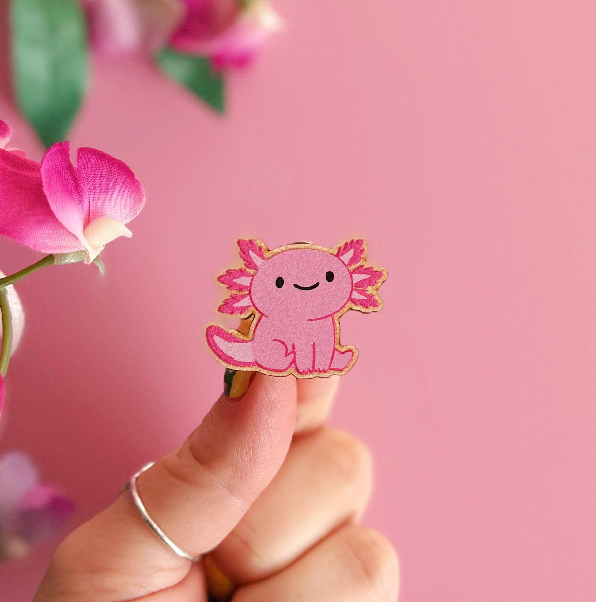 A wooden pin badge with a glossy finish, featuring an illustration of a pink axolotl.