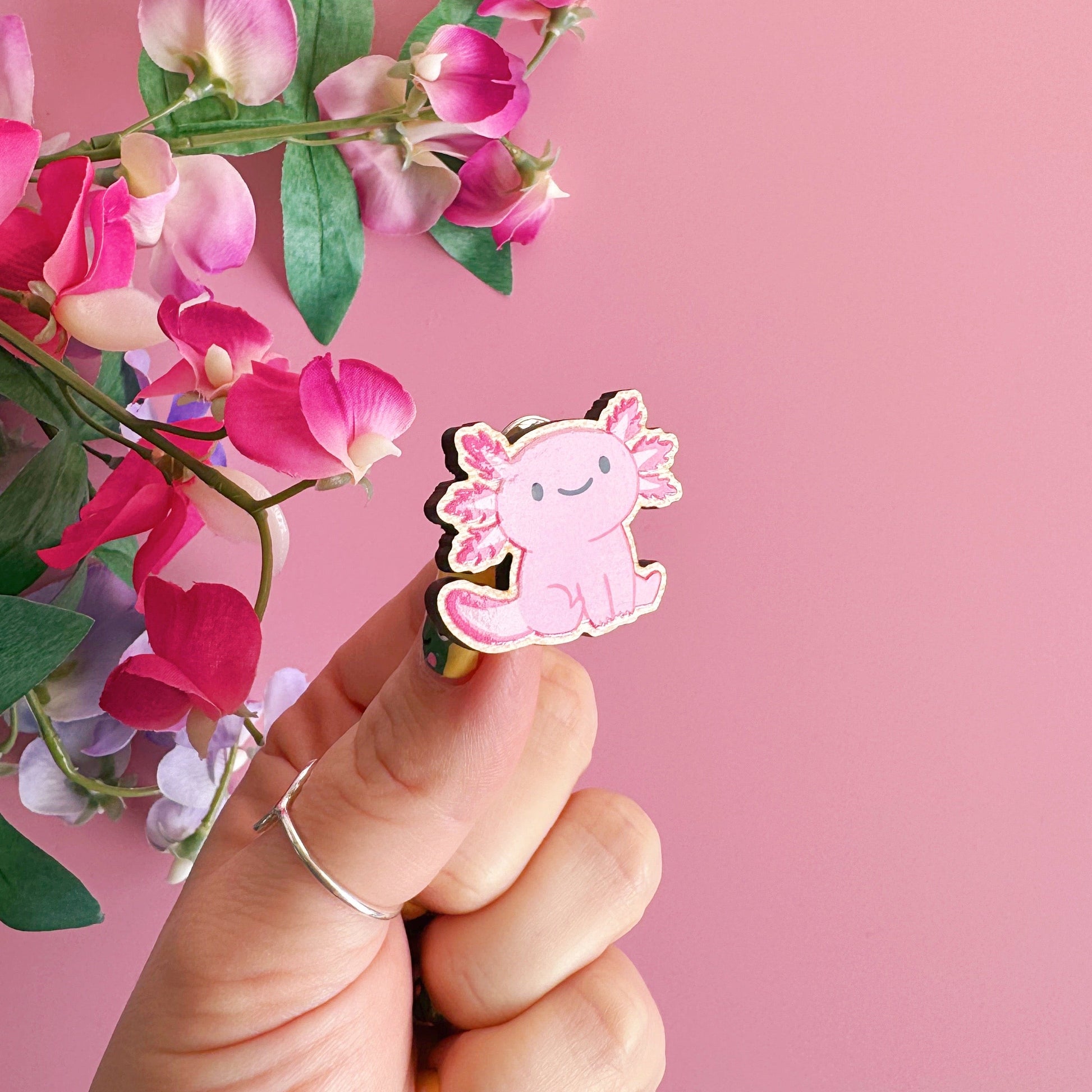 A wooden pin badge with a glossy finish, featuring an illustration of a pink axolotl.