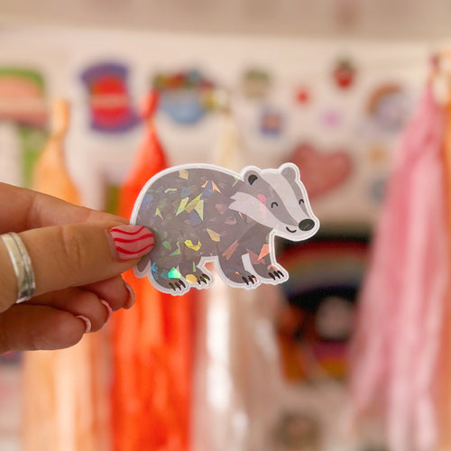 A sticker featuring a cute smiling badger on all fours, which is coated with a holographic film making it catch the sunlight.