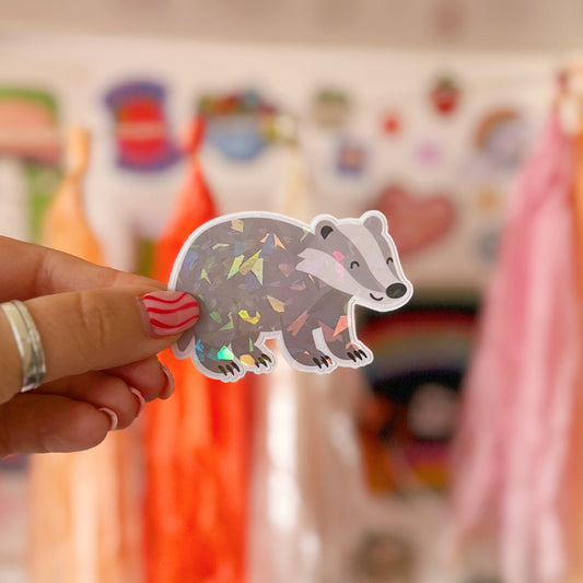 Hand holding a cute badger sticker in front of a shelf with toys and decorations.