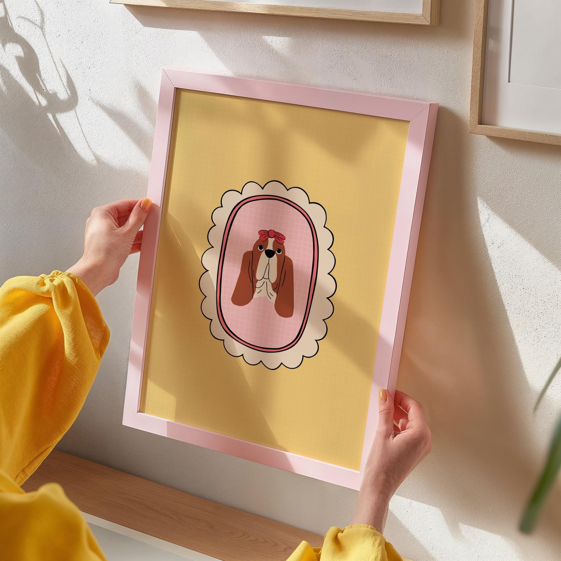 A person holding a framed art print of a Basset Hound dog with a yellow background, preparing to hang it on the wall.