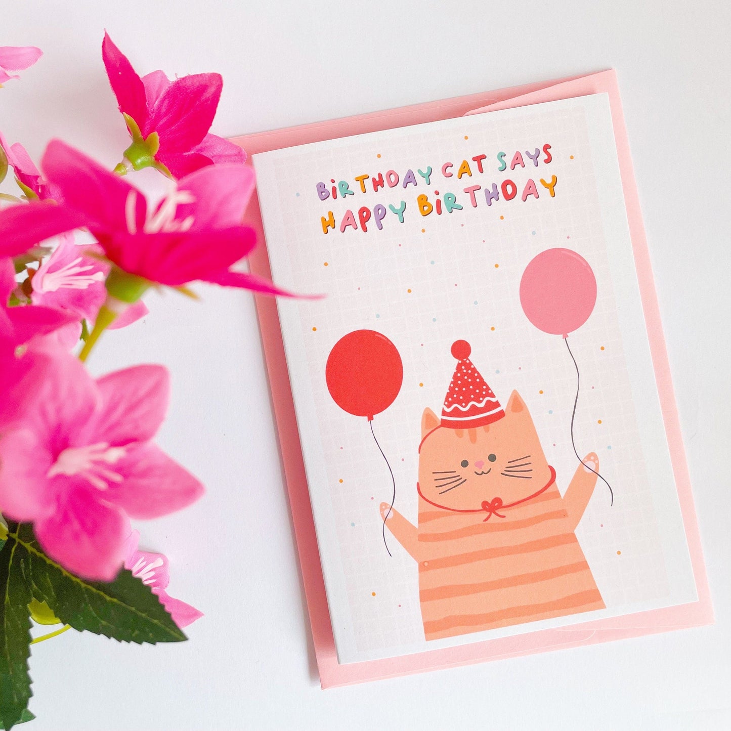 A birthday card, with a cat wearing a party hat and holding balloons. The text reads, birthday cat says happy birthday.