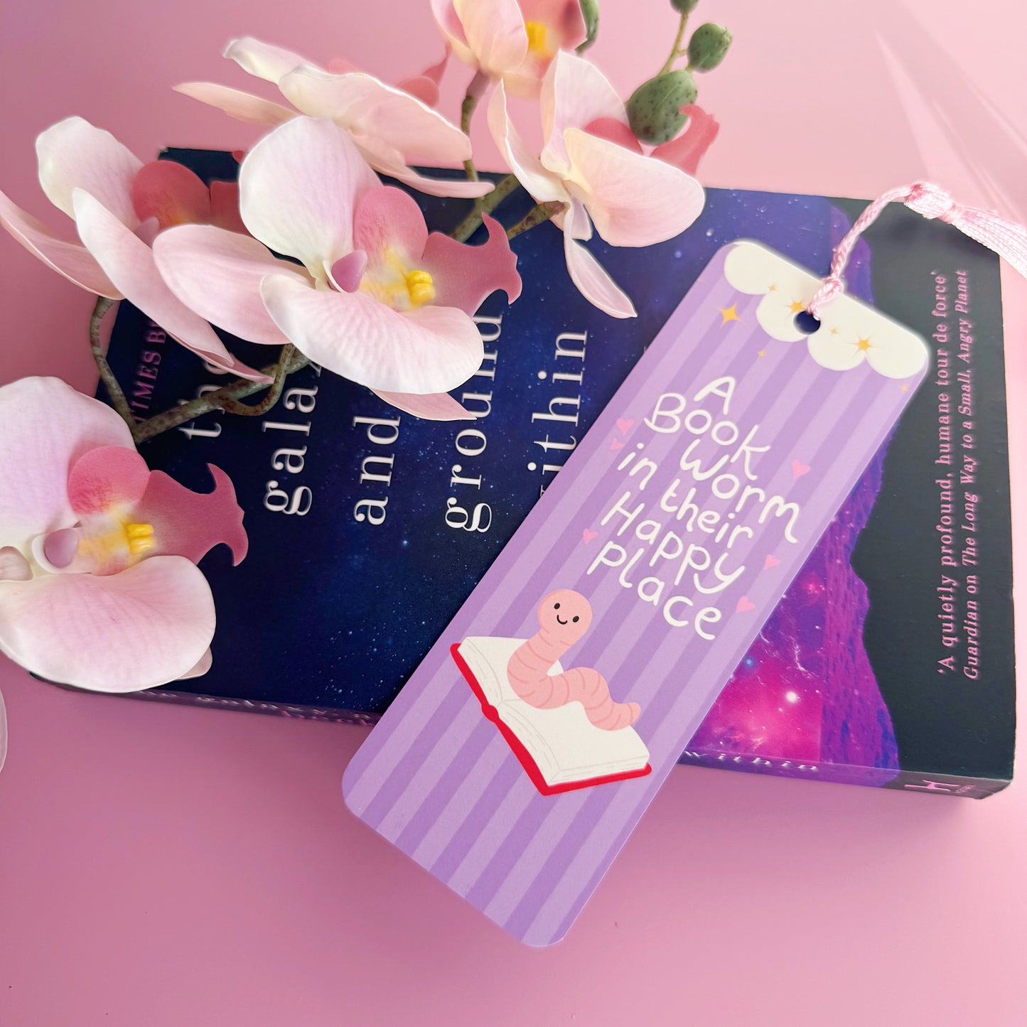 A purple bookmark with a worm illustration, and lettering that reads 'a book worm in their happy place'.