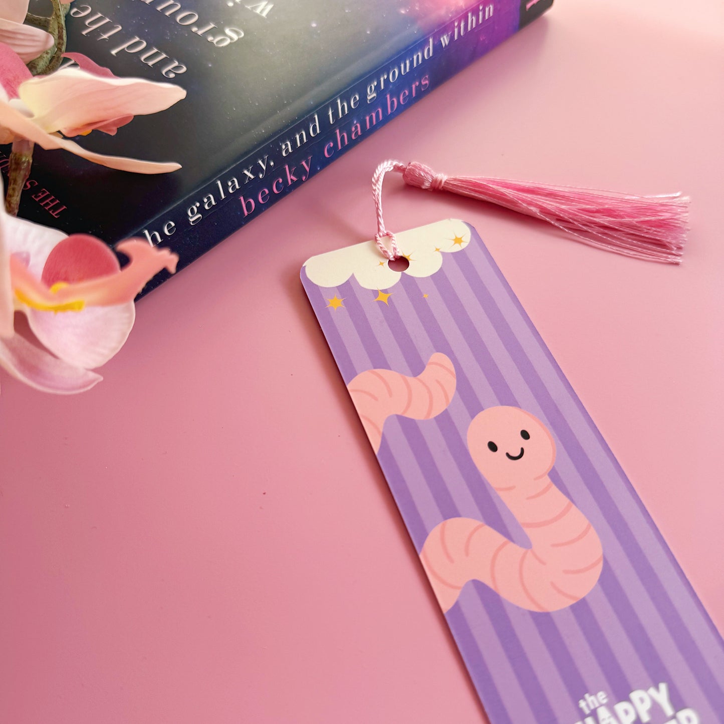 A purple bookmark with a worm illustration, and lettering that reads 'a book worm in their happy place'.