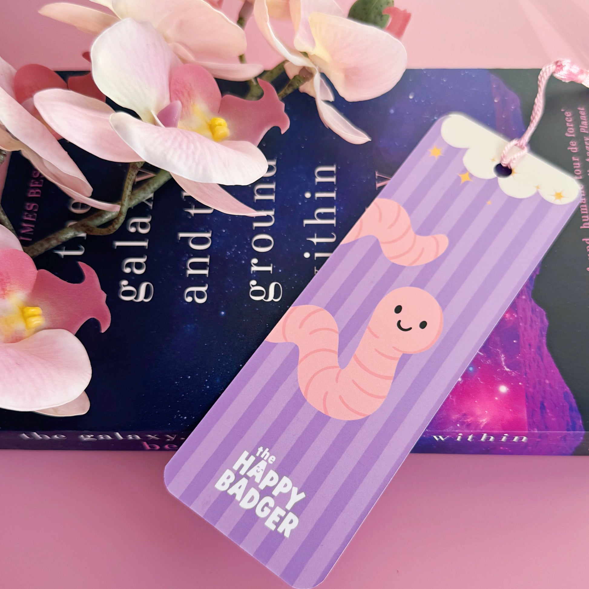 A purple bookmark with a worm illustration, and lettering that reads 'a book worm in their happy place'.