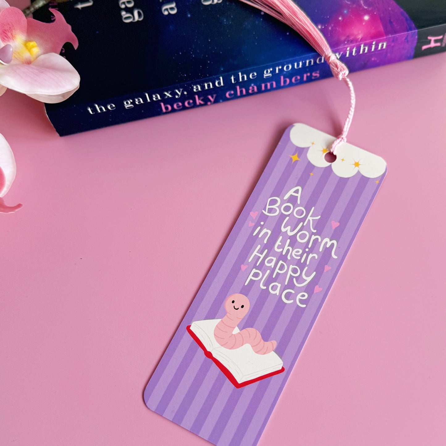 A purple bookmark with a worm illustration, and lettering that reads 'a book worm in their happy place'.