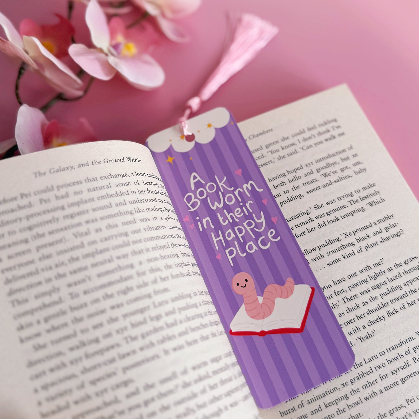 A purple bookmark with a worm illustration, and lettering that reads 'a book worm in their happy place'.