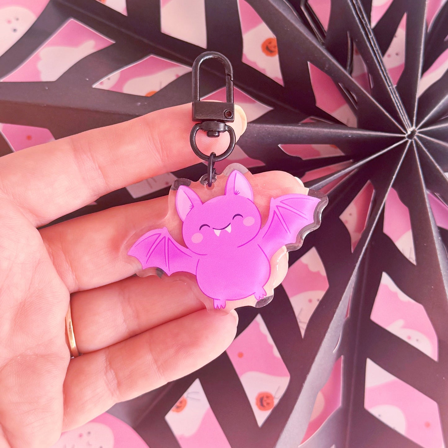 A purple acrylic bat keyring, featuring an illustration of a  cute smiling bat. The keyring has a black clasp to attach it to keys or a bag.