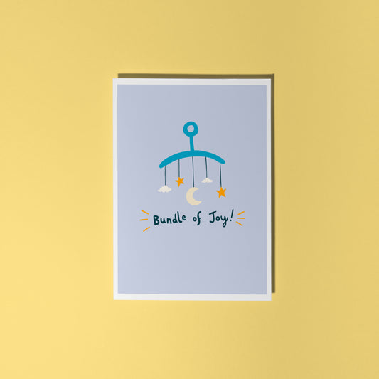 A blue greeting card with an illustration of a mobile. Underneath the illustration is hand written text that reads 'bundle of joy!'.