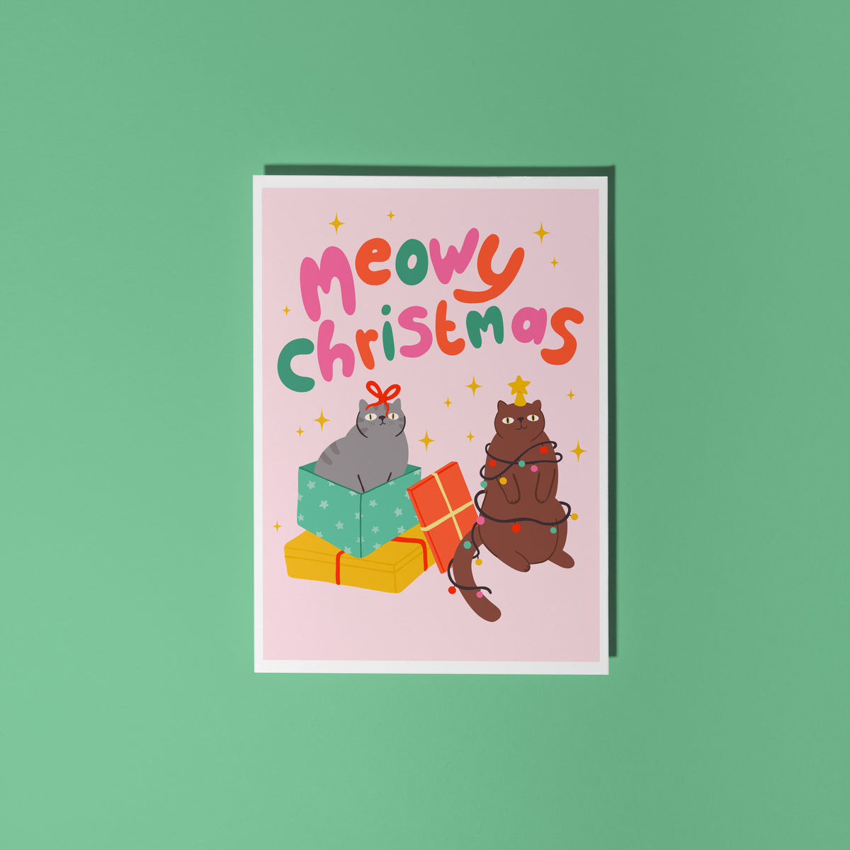 A Christmas flat lay scene showing a Christmas card with two cats on the front. One grey cat and one brown cat. The text on the card says 'Meowy Christmas' in hand lettered text.