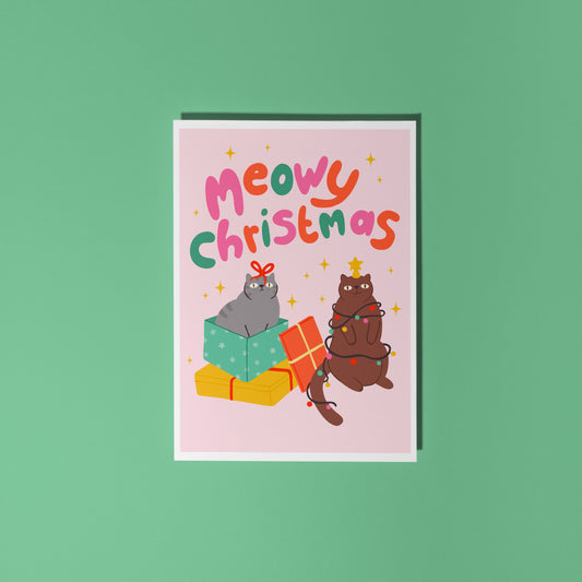 A Christmas flat lay scene showing a Christmas card with two cats on the front. One grey cat and one brown cat. The text on the card says 'Meowy Christmas' in hand lettered text.