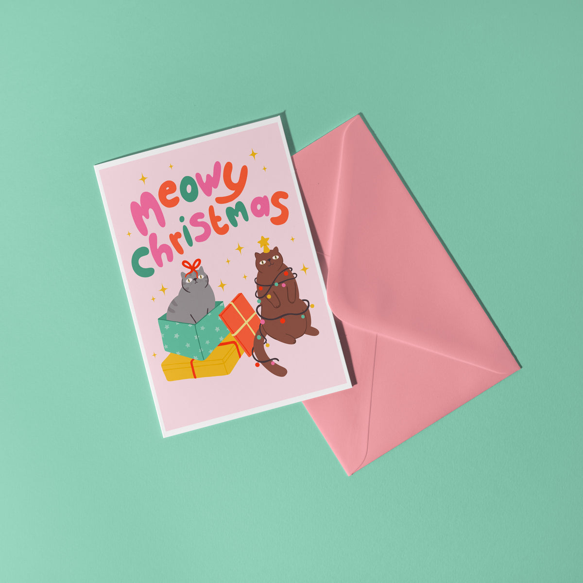 A Christmas flat lay scene showing a Christmas card with two cats on the front. One grey cat and one brown cat. The text on the card says 'Meowy Christmas' in hand lettered text.