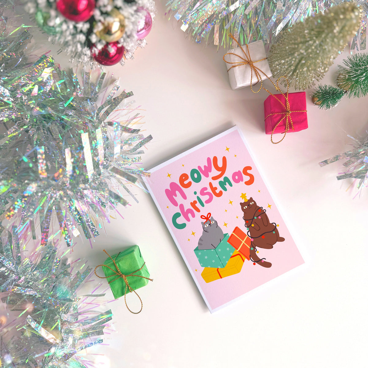A Christmas flat lay scene showing a Christmas card with two cats on the front. One grey cat and one brown cat. The text on the card says 'Meowy Christmas' in hand lettered text.