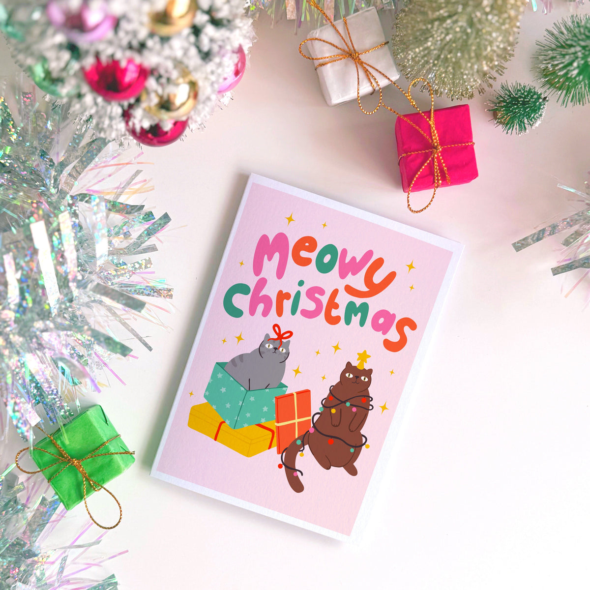 A Christmas flat lay scene showing a Christmas card with two cats on the front. One grey cat and one brown cat. The text on the card says 'Meowy Christmas' in hand lettered text.