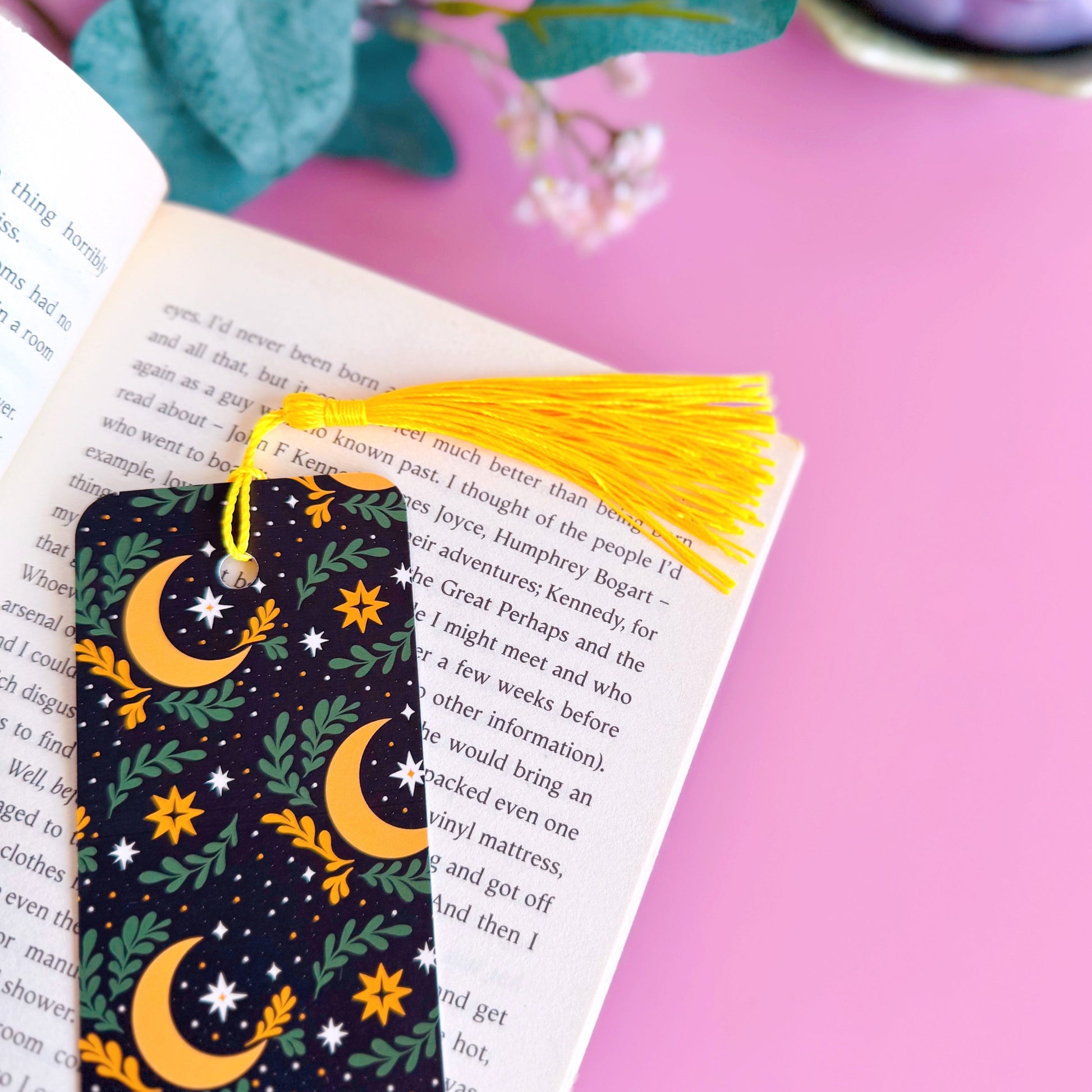 A black bookmark, with a yellow and green celestial themed pattern. It features large moons, green fern leaves and little yellow and white stars. The bookmark has a coordinating tassel in yellow.