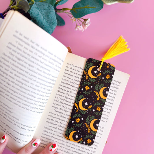 A black bookmark, with a yellow and green celestial themed pattern. It features large moons, green fern leaves and little yellow and white stars. The bookmark has a coordinating tassel in yellow.