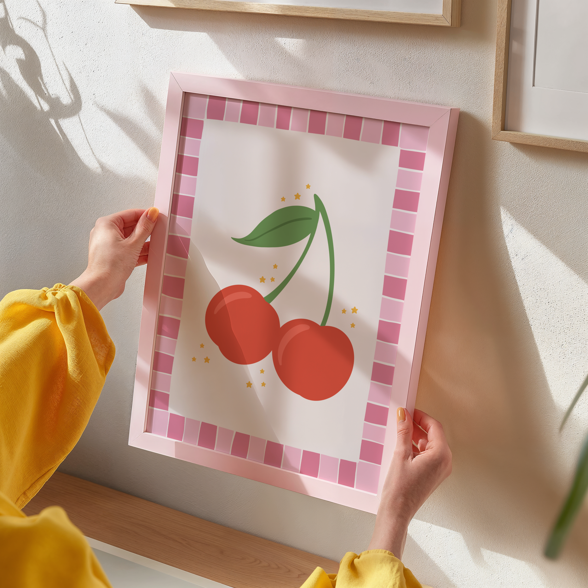 A print with an illustration of a pair of cherries, on a soft pink background surrounded by a bright pink hand drawn border.