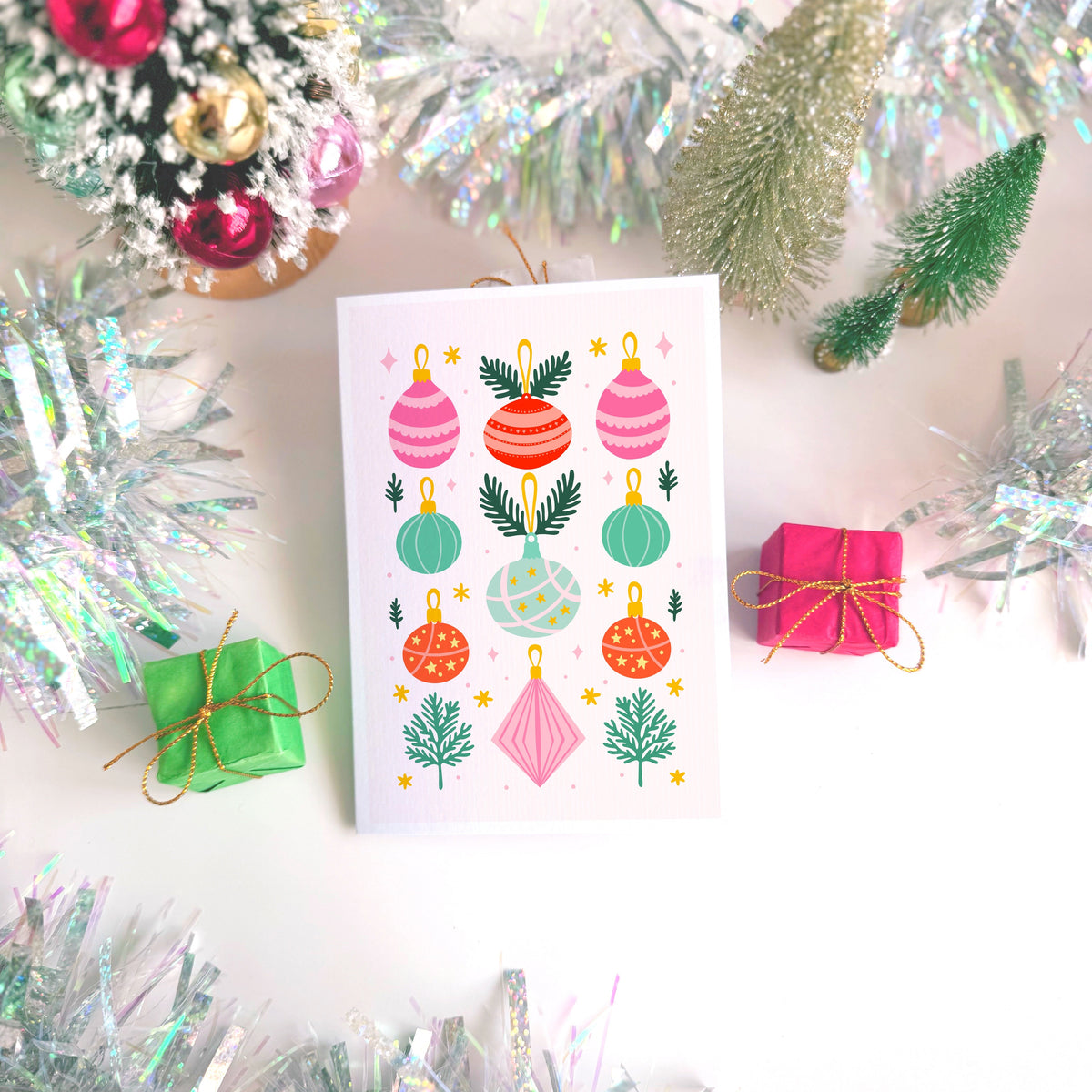 A white Christmas ornament Christmas card featuring colourful illustrations of baubles, pine trees, and stars, styled on a white background with festive tinsel, miniature gifts, and a decorated Christmas tree.