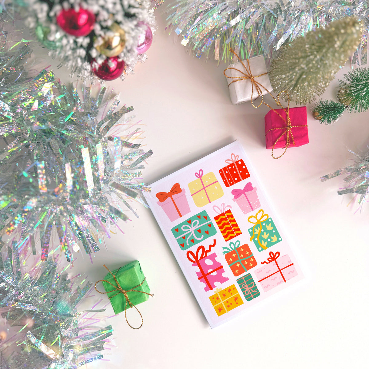 A Christmas card featuring colourful gift boxes and bows, styled on a light background with festive tinsel, miniature wrapped presents, and a small decorated tree.