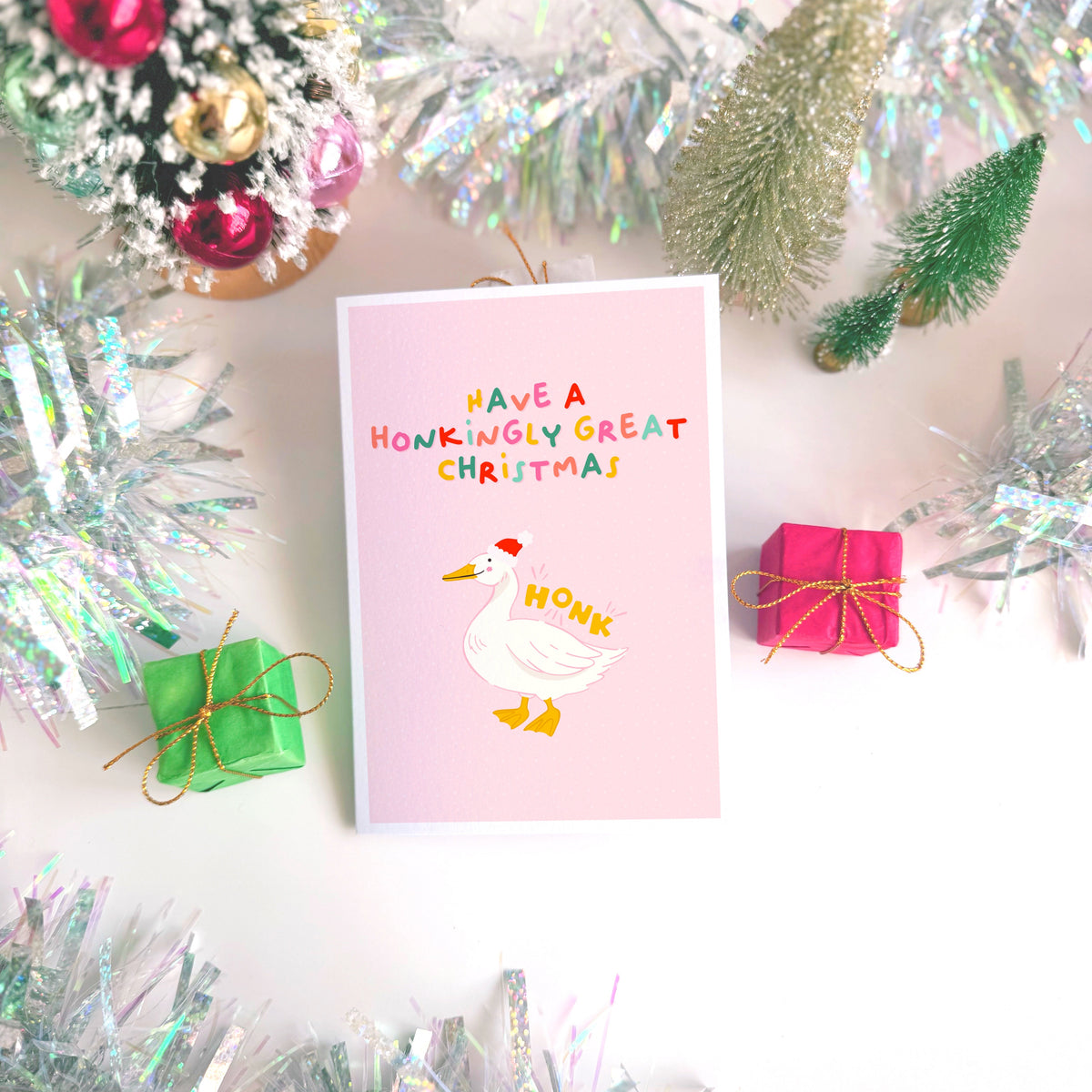 A pink Christmas card with an illustration of a goose with a red Santa hat on. The multicoloured text above the illustration reads 'have a honkingly great Christmas'.
