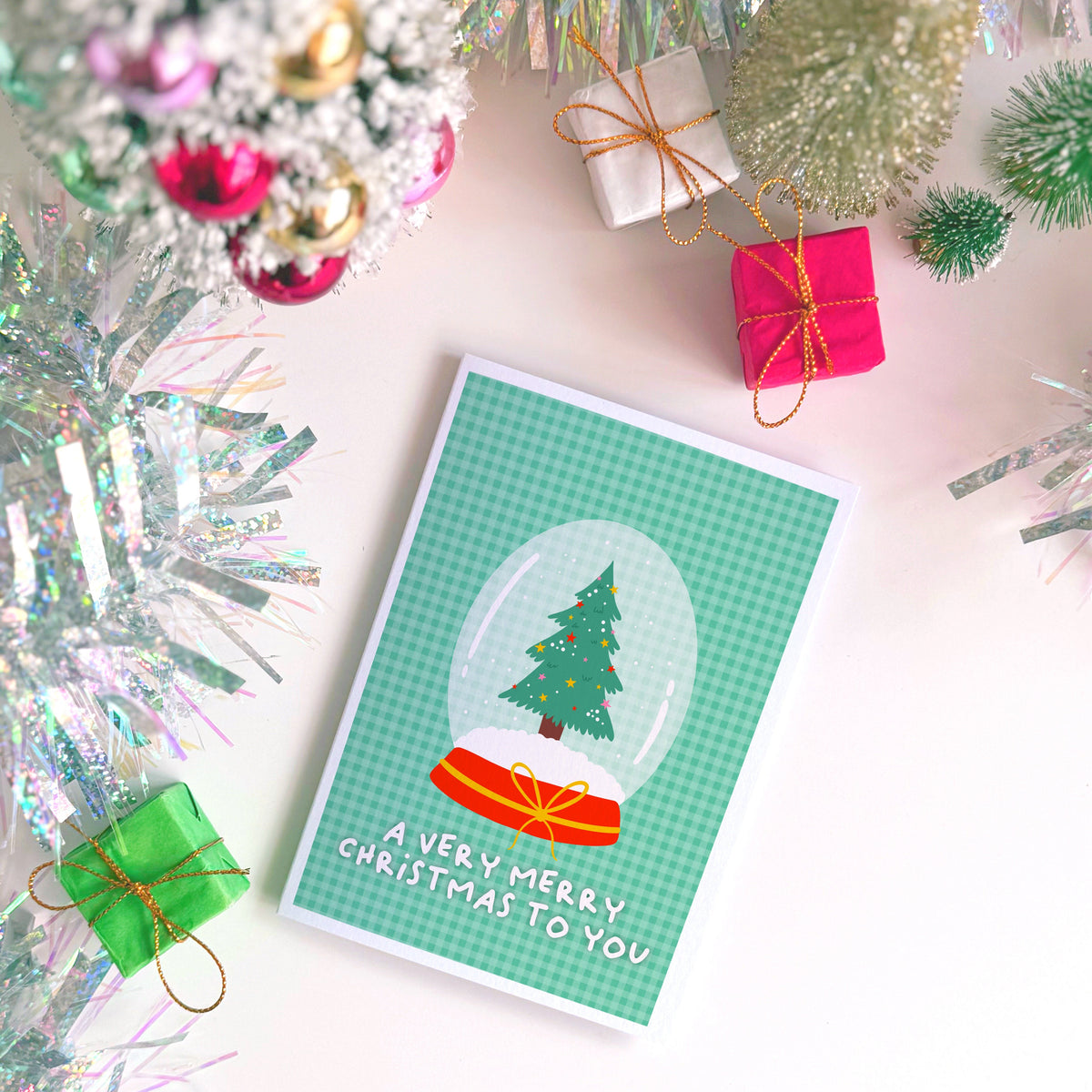 A festive Christmas greeting card with a green gingham background, featuring a snow globe illustration with a Christmas tree and the text 'A VERY MERRY CHRISTMAS TO YOU'. The card is styled in a flat-lay arrangement with silver tinsel, miniature gift boxes, and a small artificial tree on a white surface.