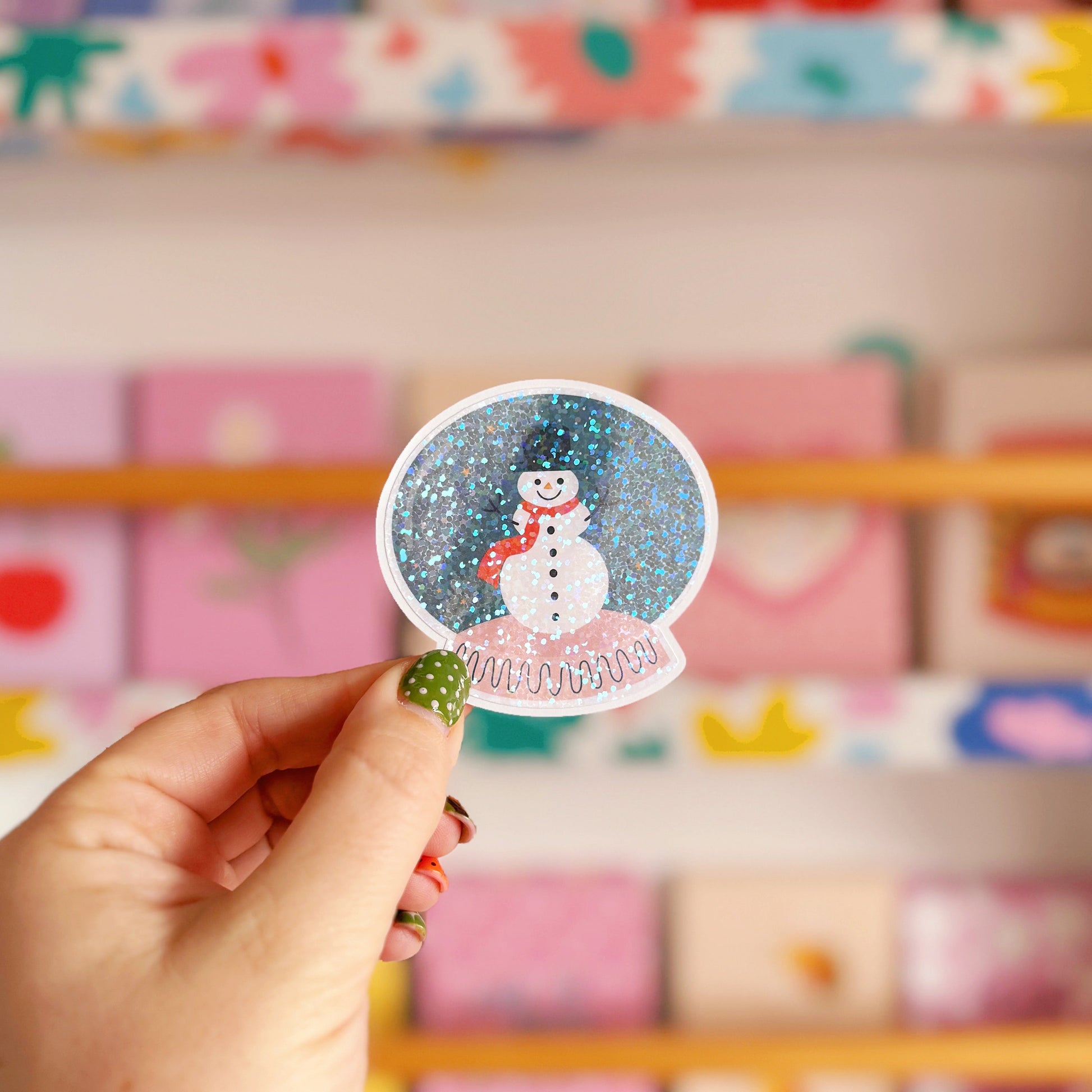 Hand holding a snow globe sticker with a snowman in front of a colorful shelf.