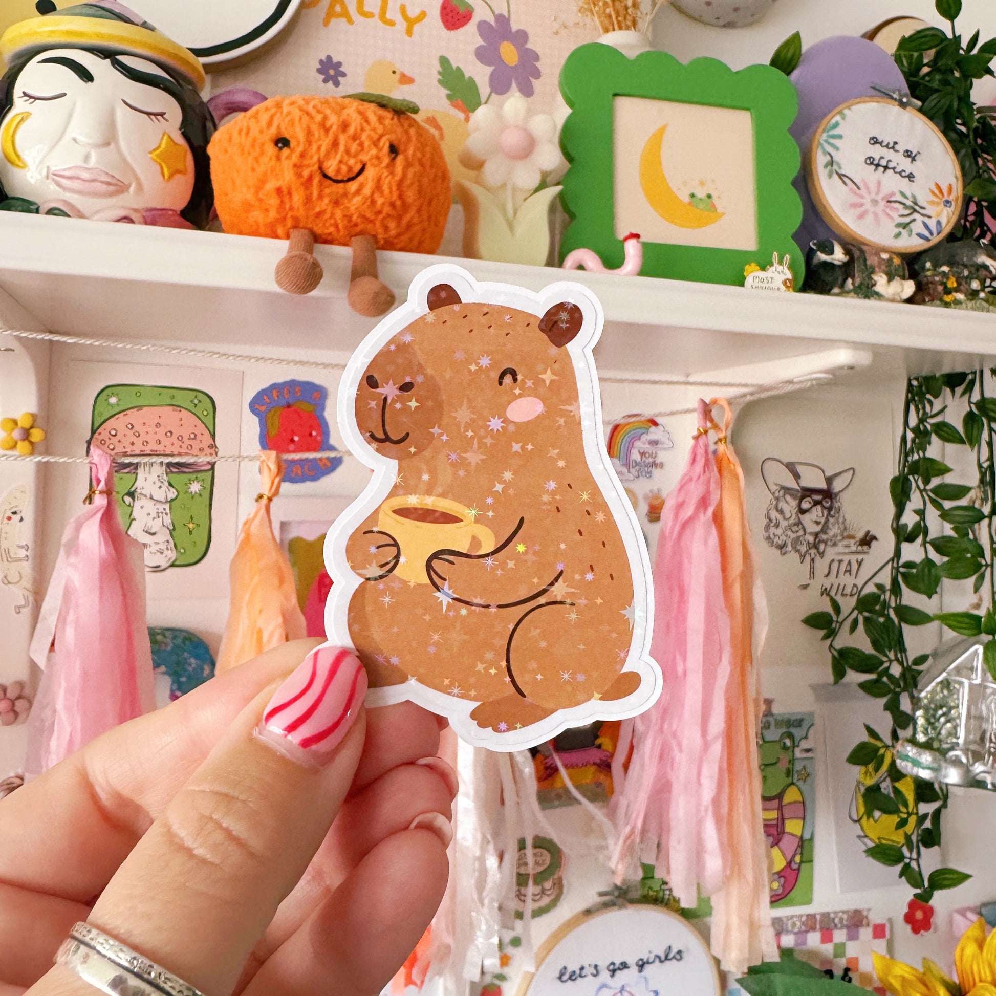 A sticker featuring a cute smiling capybara, holding a hot mug of tea or coffee. The sticker is coated with a starry holographic film.