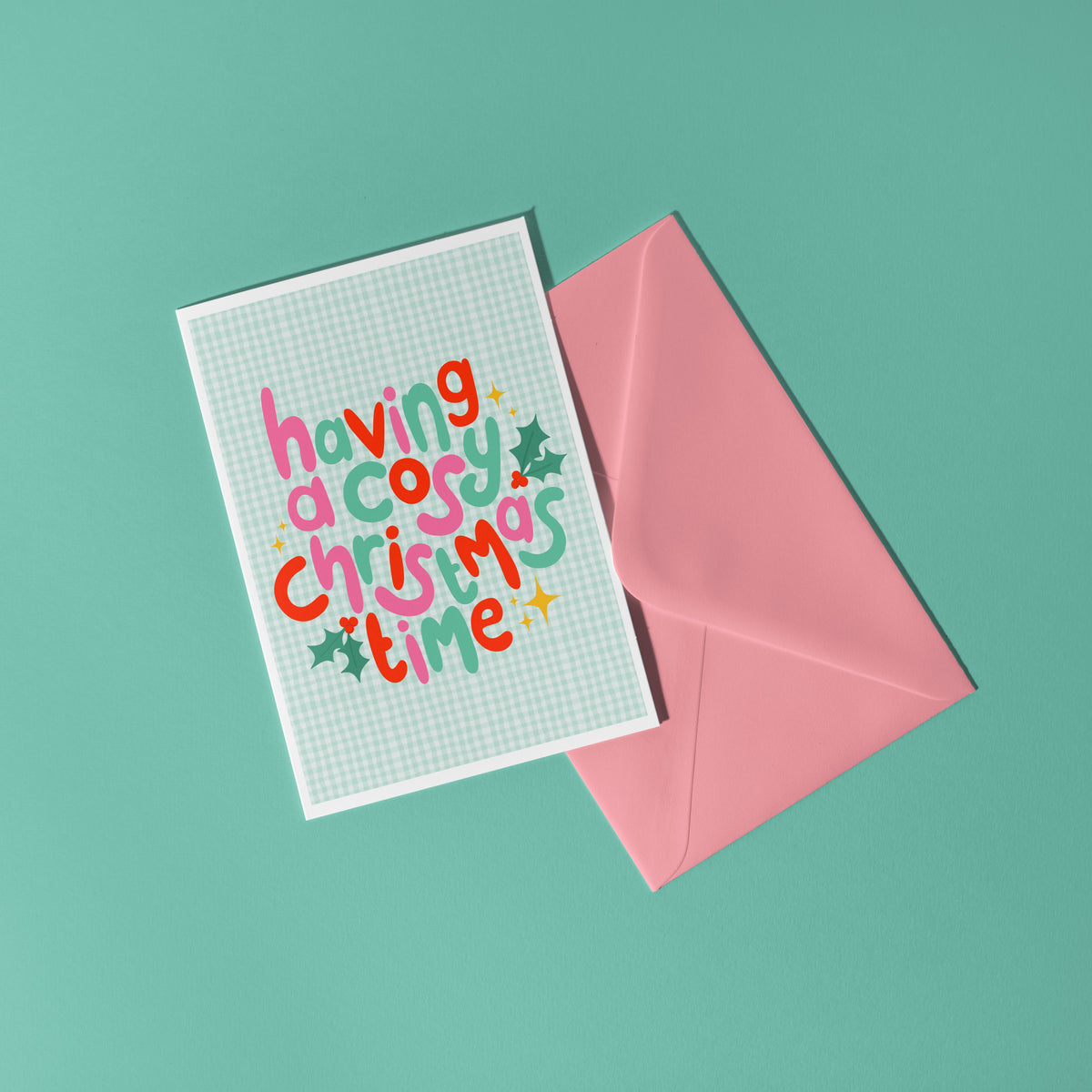 A square greeting card with colourful playful text reading 'having a cosy Christmas time' on a pale green gingham background, shown with a pink envelope on a mint green surface.