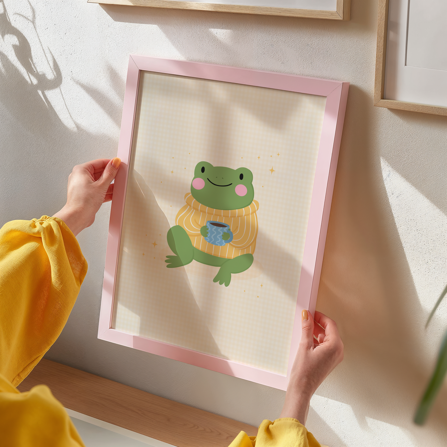 Framed artwork of a frog wearing a sweater with a cup on a pale yellow gingham background.