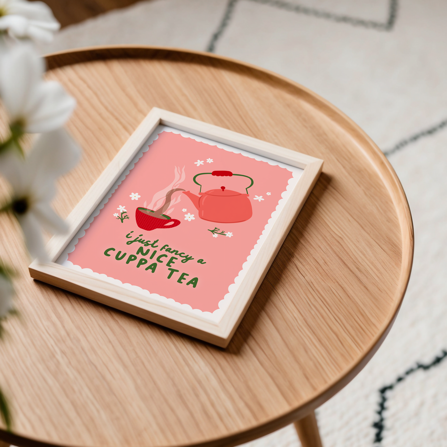 A print with a pink background, with a beige scalloped border, with an illustration of a vintage style kettle and a mug of tea. the quote at the bottom reads 'I just fancy a nice cuppa tea'