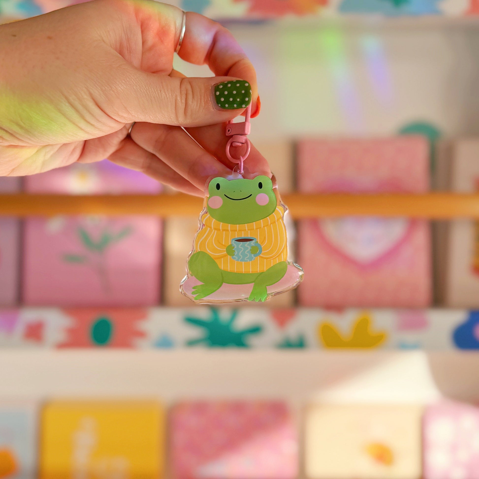 Hand holding a keychain with a cartoon frog design in front of a wall of colourful postcards.