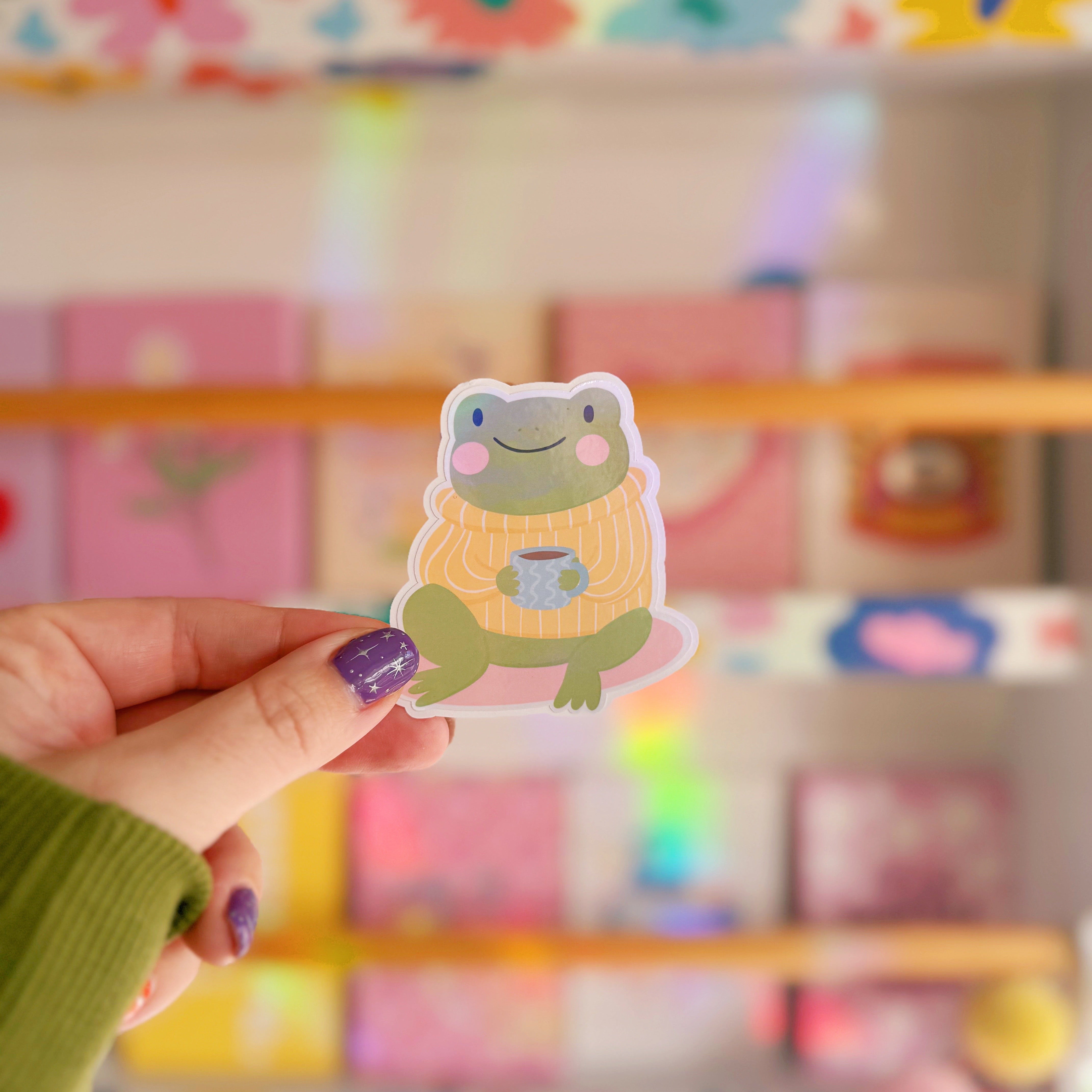 Hand holding a cartoon frog sticker with a colorful background