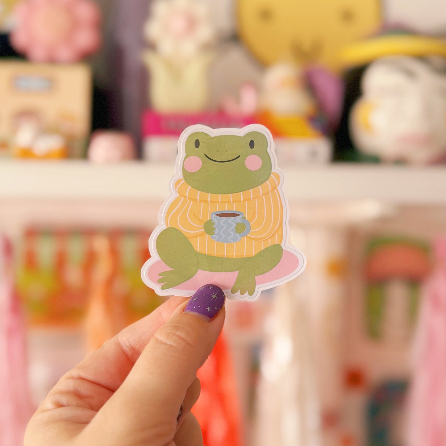A sticker of a cute little frog, sitting down wearing a yellow striped jumper. The frog is also holding a mug of coffee/tea.