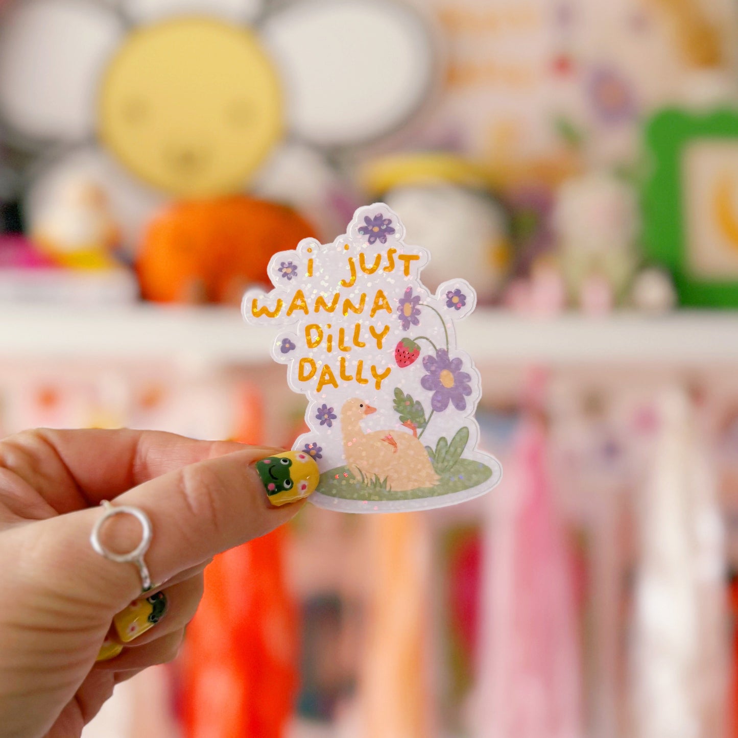 A cute holographic sticker with a duck laying on it's back, next to purple flowers. Above it are the words 'I Just Wanna Dilly Dally' in yellow.