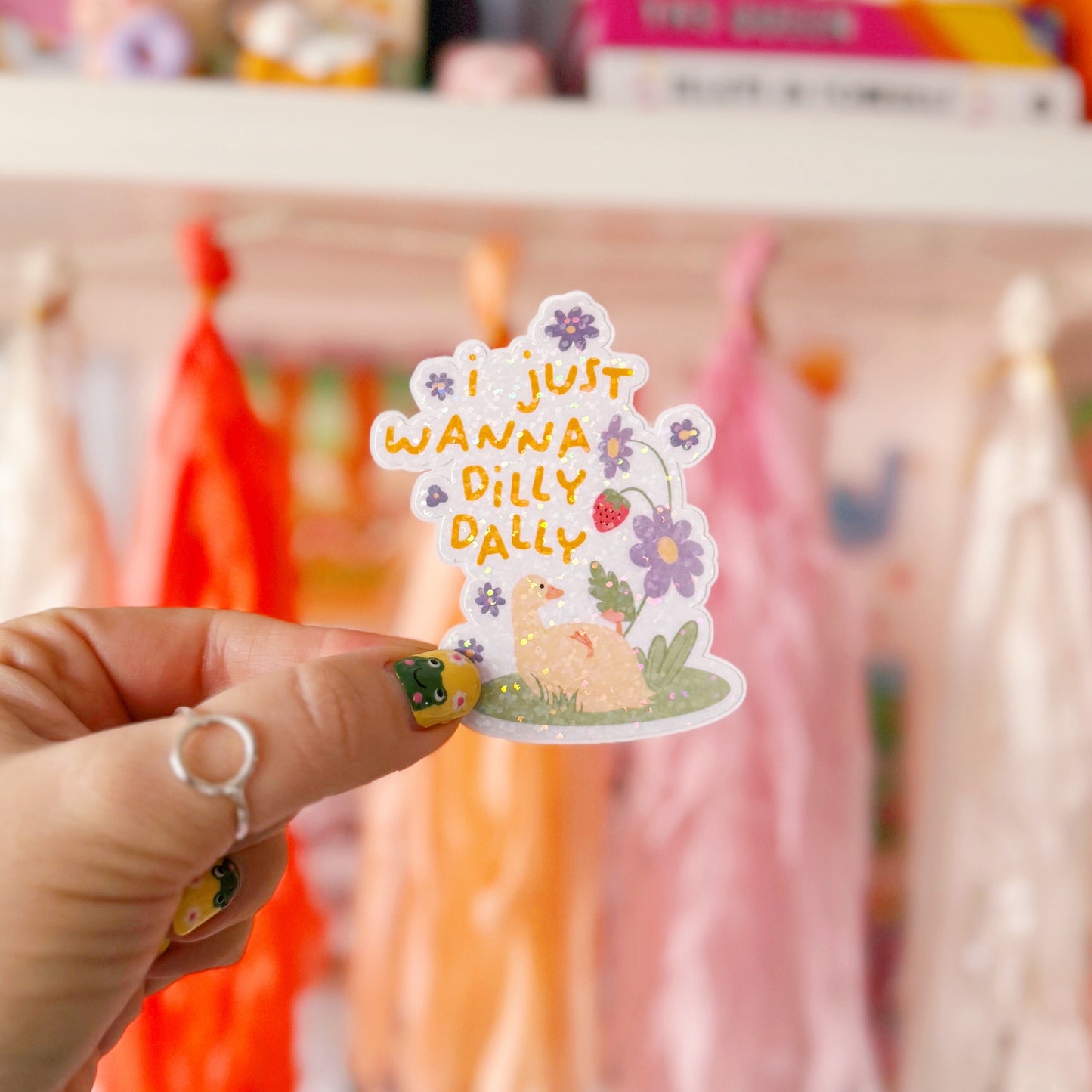 A cute holographic sticker with a duck laying on it's back, next to purple flowers. Above it are the words 'I Just Wanna Dilly Dally' in yellow.