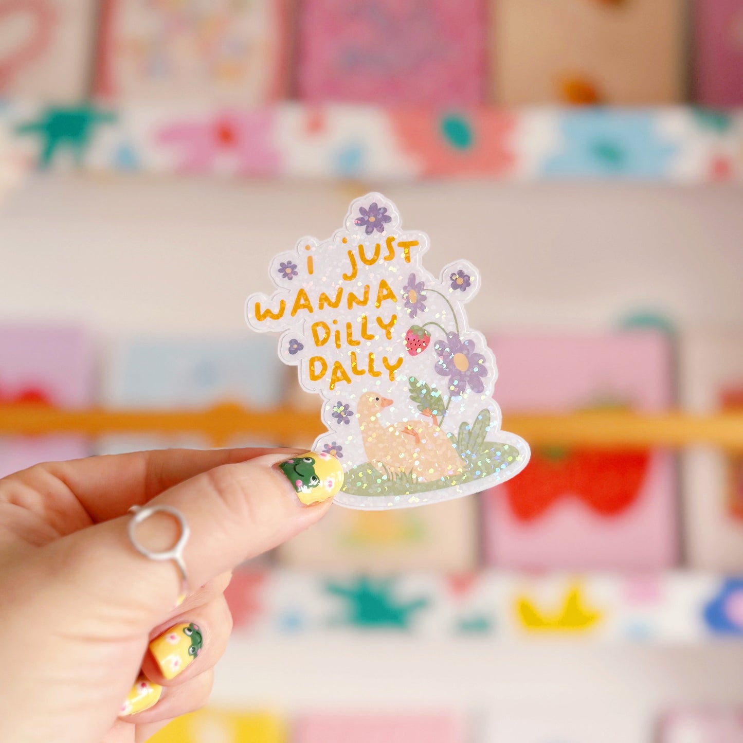 A cute holographic sticker with a duck laying on it's back, next to purple flowers. Above it are the words 'I Just Wanna Dilly Dally' in yellow.