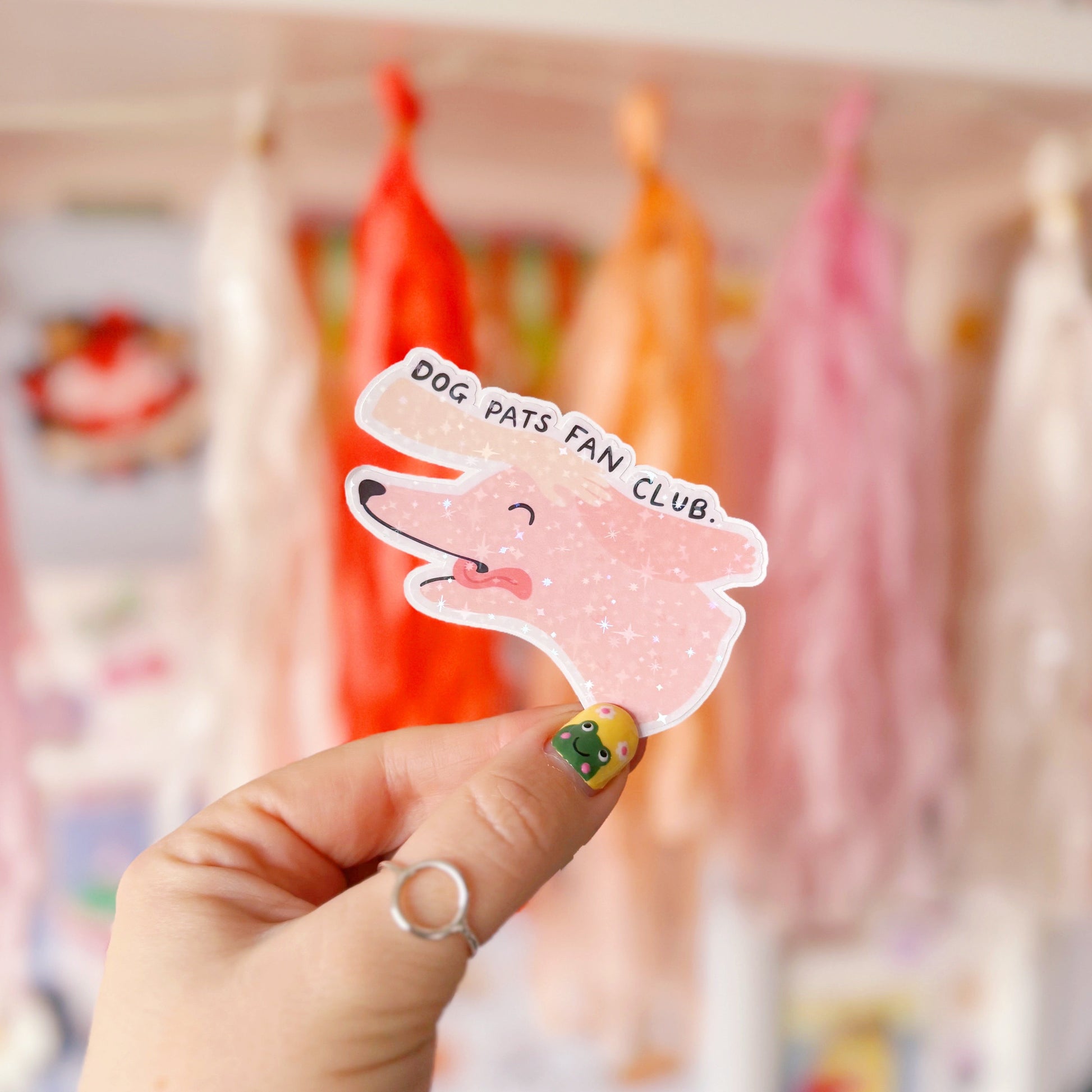A holographic sticker of a pink dog, with its tongue sticking out, with a hand patting its head. The text reads 'Dog Pats Fan Club.'