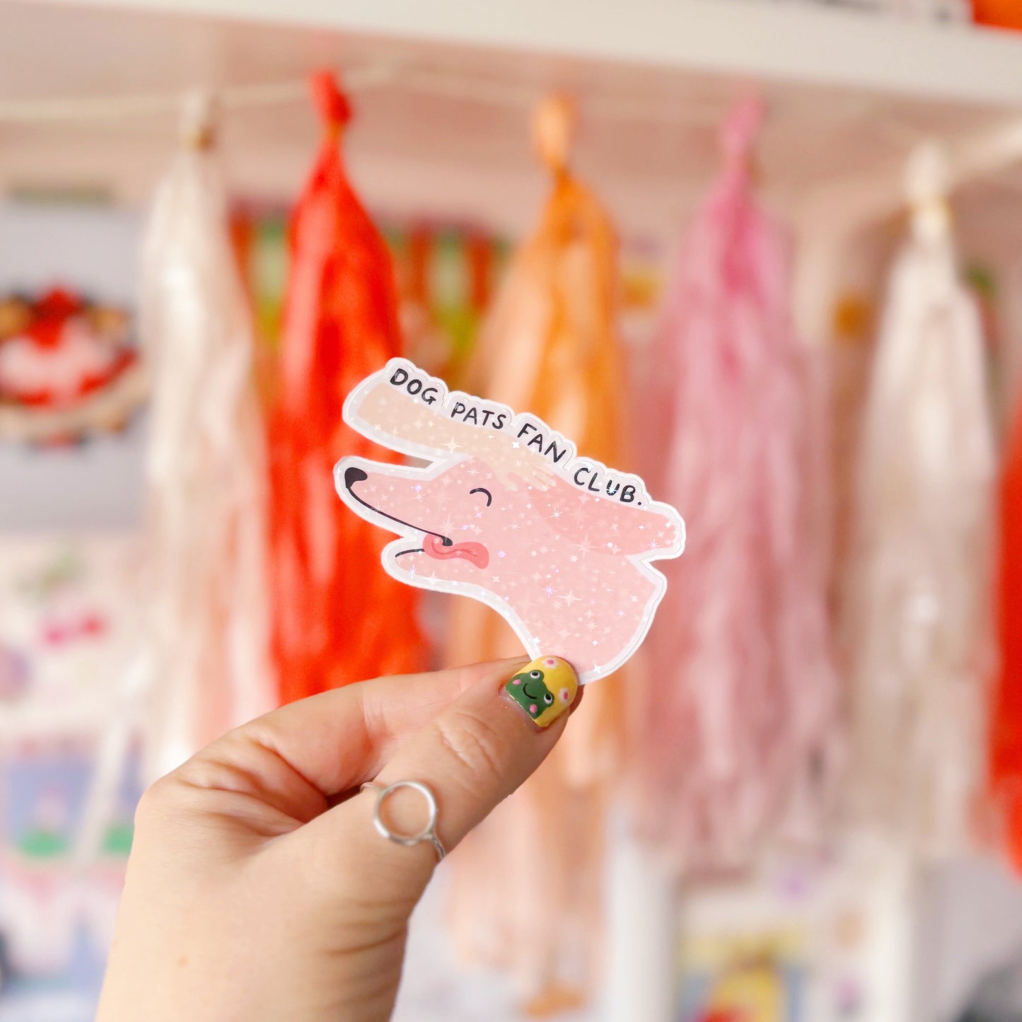 A holographic sticker of a pink dog, with its tongue sticking out, with a hand patting its head. The text reads 'Dog Pats Fan Club.'