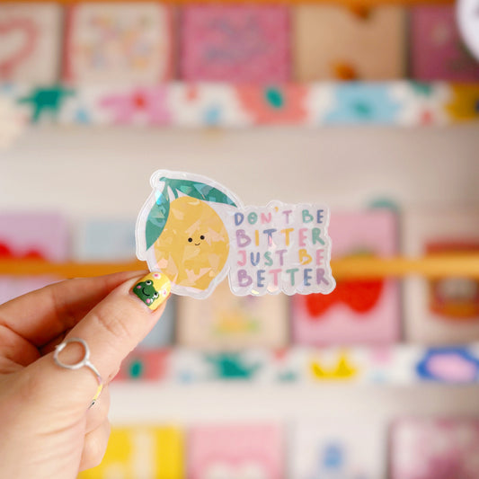 A holographic sticker, of a smiling lemon and the quote don't be bitter just be better. The sticker has an easy peel lip.