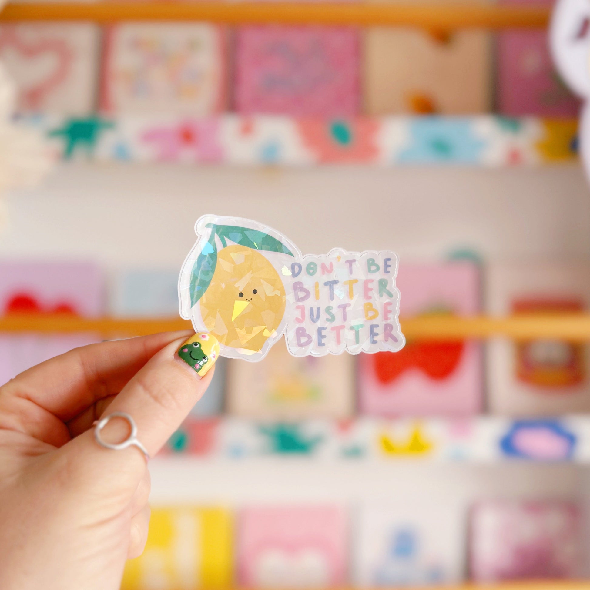 A holographic sticker, of a smiling lemon and the quote don't be bitter just be better. The sticker has an easy peel lip.