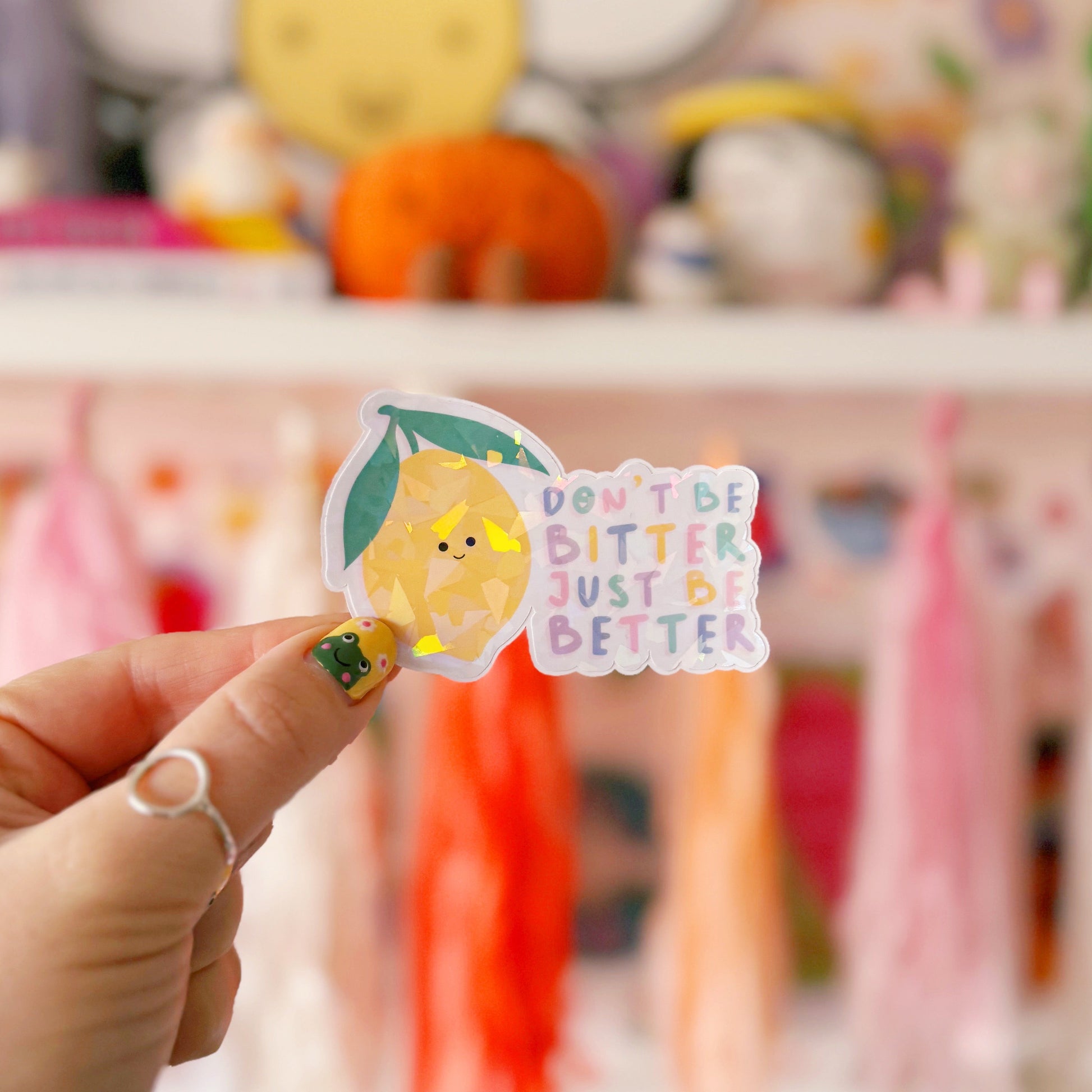 A holographic sticker, of a smiling lemon and the quote don't be bitter just be better. The sticker has an easy peel lip.