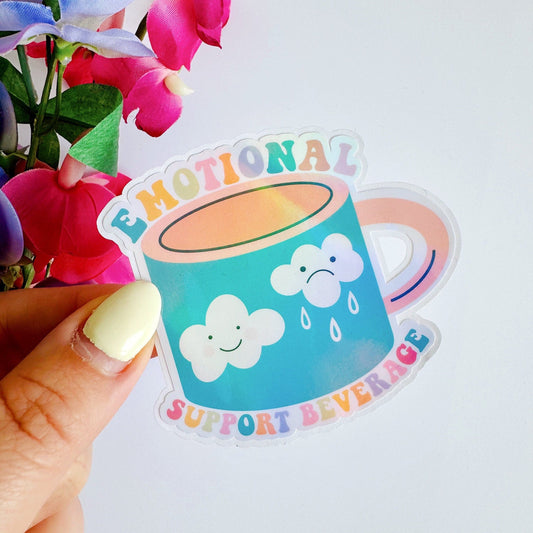 A holographic sticker, of a mug with a happy and a sad cloud on the front and the words emotional support beverage around the mug. The sticker has an easy peel lip.