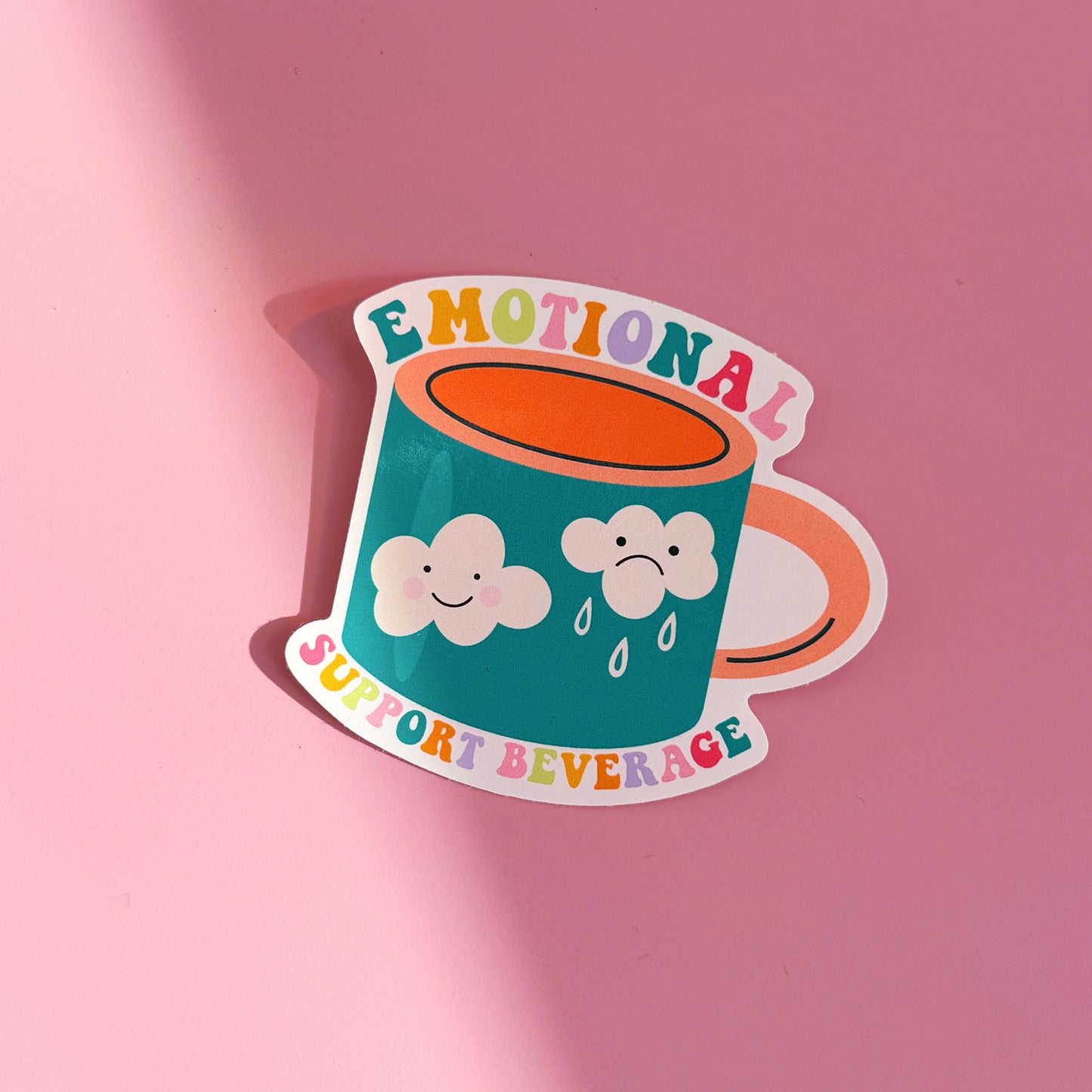 A waterproof sticker, featuring an illustration of a mug with a sad and a happy cloud on the front. Around the mug are the words 'emotional support beverage'.